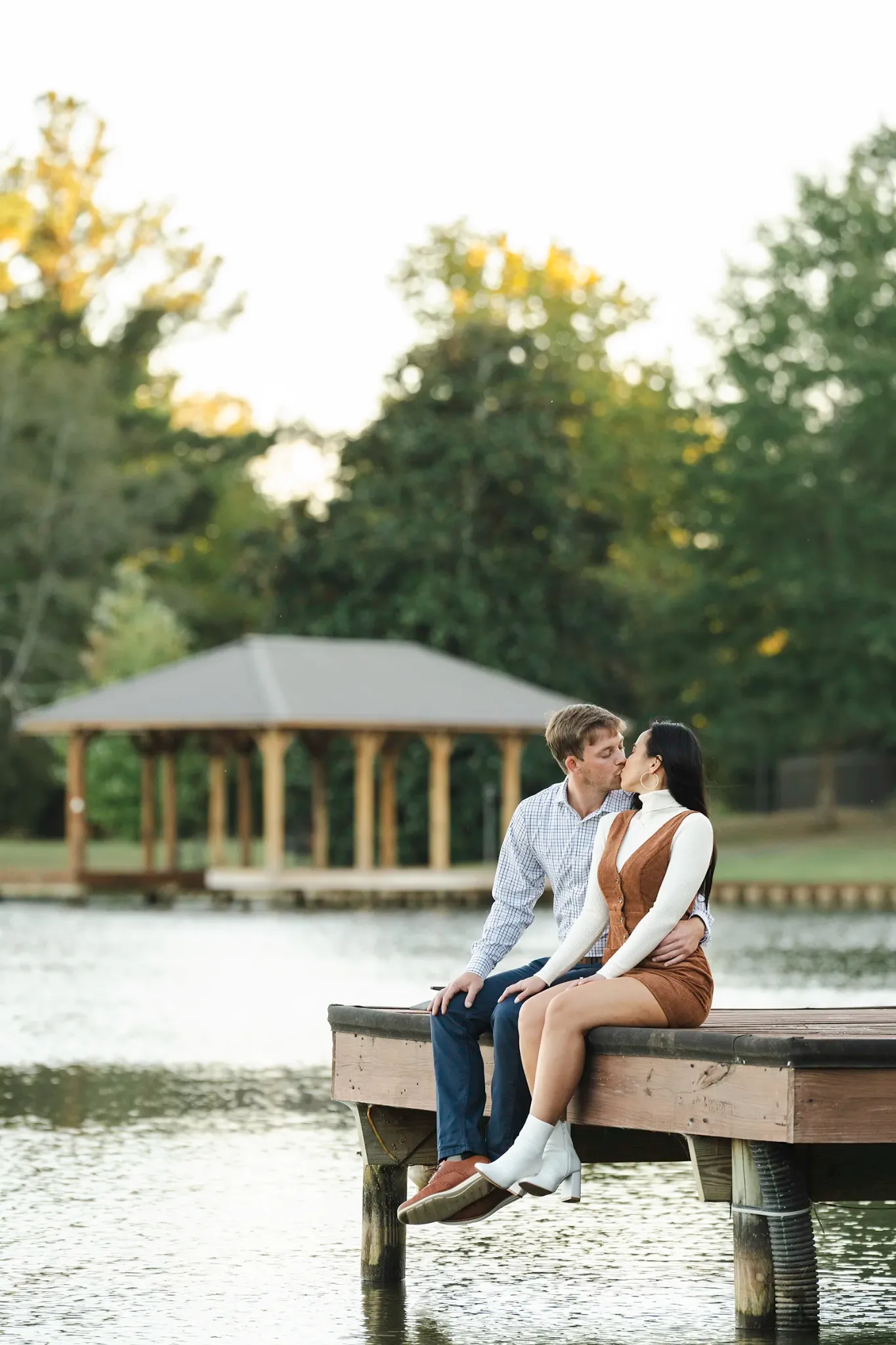 Engaged couple kissing while seated on a dock beside a lake