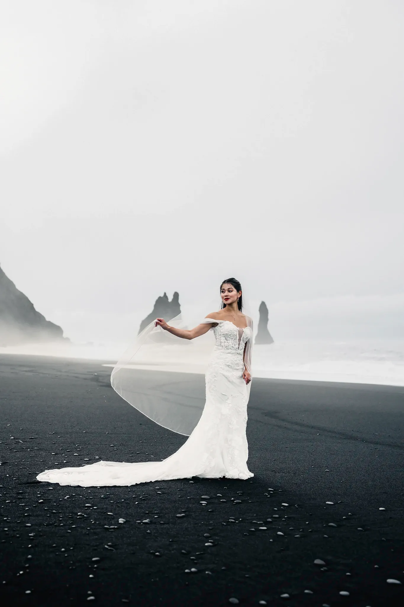 Bride in a fitted gown standing on a black-sand beach in misty coastal scenery
